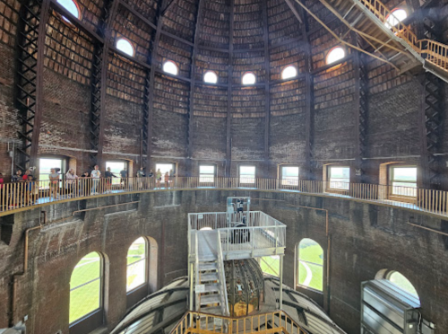 Interior view of a large, circular structure with wooden beams, a spiral staircase, and arched windows. Visitors are present.