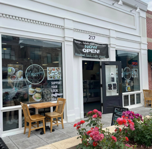 Exterior of a café with a "NOW OPEN!" sign, featuring outdoor seating and colorful flowers in front.