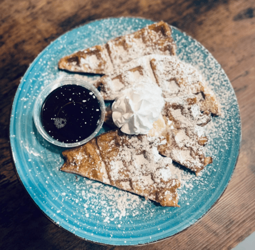 A blue plate with sliced waffles dusted with powdered sugar, topped with whipped cream and a side of syrup.