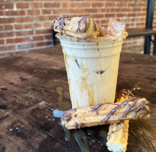 A cup of iced drink topped with chocolate, surrounded by chocolate-drizzled churros on a wooden table.