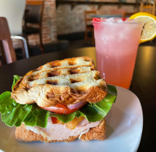 A toasted sandwich with turkey, lettuce, and tomato on a plate, alongside a pink drink with a lemon slice.