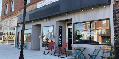 Exterior of a cozy café with a "Welcome" sign, outdoor seating, and an "Open" sign in the window.