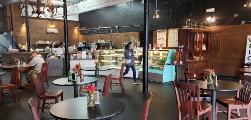 A cozy café interior with wooden tables, chairs, and a display counter, featuring flowers on tables and a customer at the counter.
