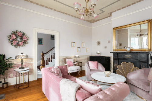 A stylish living room with pink furniture, a large mirror, floral decor, and elegant lighting.