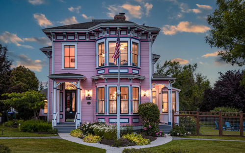 A charming pink Victorian house with a flag, surrounded by lush greenery and a clear blue sky at dusk.