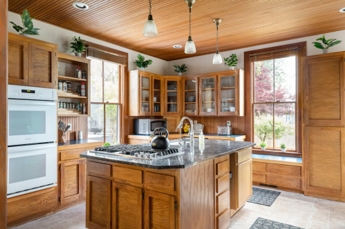 Bright kitchen with wooden cabinets, a central island, and large windows, featuring modern appliances and plants.