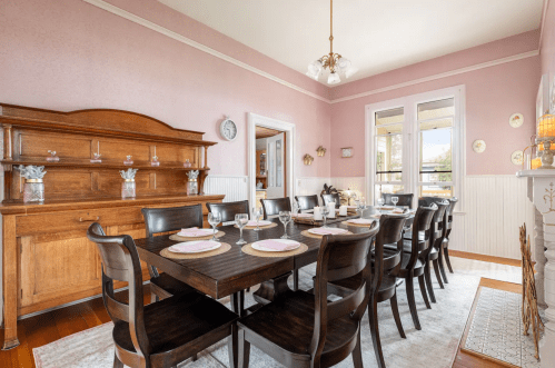 A spacious dining room with a long wooden table set for dinner, surrounded by chairs and decorated in soft pink tones.