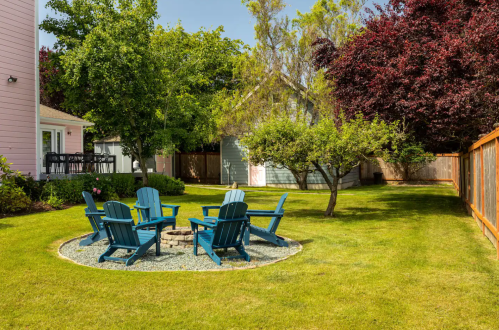 A sunny backyard with a circular fire pit surrounded by blue chairs, lush green grass, and trees.