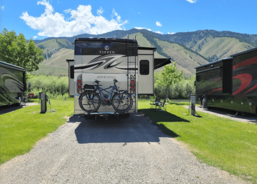 A parked RV with bicycles on a rack, surrounded by green grass and mountains under a blue sky.