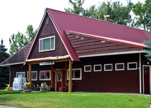 A brown wooden building with a red metal roof, featuring a sign for ice and surrounded by greenery.