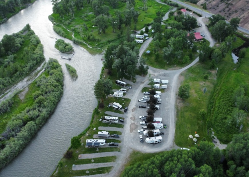 Aerial view of a campground with RVs near a winding river, surrounded by greenery and trees.