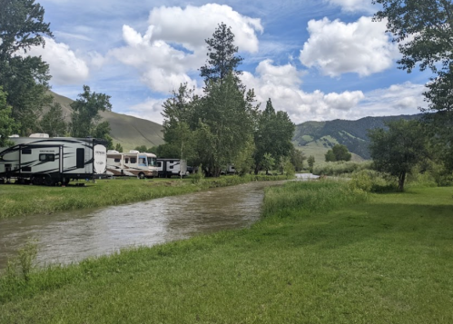 A serene landscape featuring RVs parked by a river, surrounded by lush greenery and mountains under a partly cloudy sky.