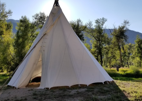 A white teepee stands in a green landscape, surrounded by trees and mountains under a bright sky.