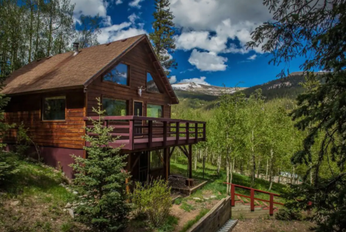 A wooden cabin surrounded by trees, with a deck overlooking a mountainous landscape under a blue sky with clouds.