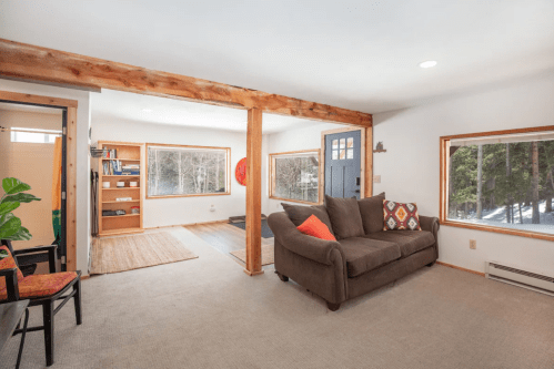 Cozy living room with a brown couch, wooden beams, and large windows overlooking a snowy landscape.