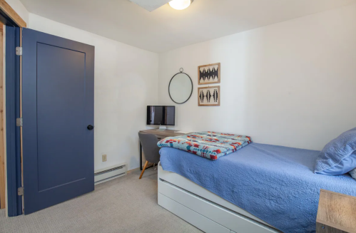 Cozy bedroom with a blue bedspread, a desk, a TV, and wall art, featuring a blue door and neutral walls.