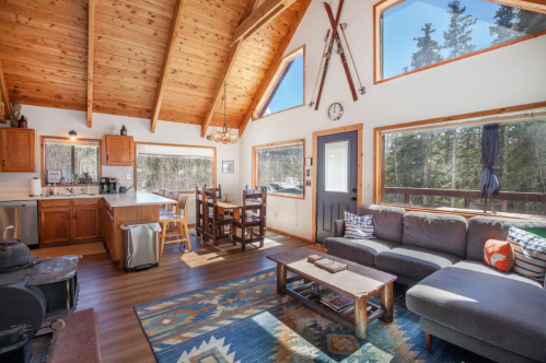 Cozy cabin interior with wooden beams, large windows, a sofa, dining area, and rustic decor.