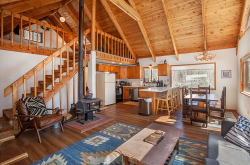 Cozy cabin interior with wooden beams, a staircase, dining area, and a wood stove, surrounded by large windows.