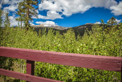 A wooden railing in the foreground with lush greenery and mountains under a blue sky with fluffy clouds in the background.