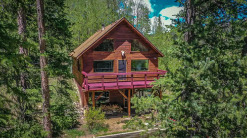 A wooden cabin surrounded by tall trees, featuring a deck and large windows, set in a lush, green landscape.