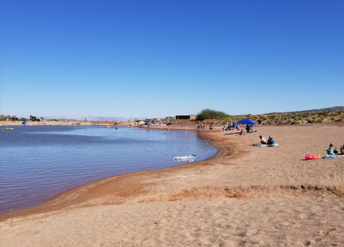 A sandy beach by a calm lake with people enjoying the sun and water activities under a clear blue sky.