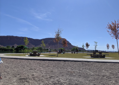 A park with sandy ground, picnic tables, and trees, set against a backdrop of a rocky mountain under a clear blue sky.