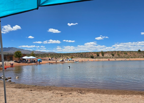 A sunny beach scene with people enjoying the water, umbrellas, and mountains in the background under a blue sky.
