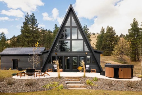 A modern A-frame cabin with large windows, surrounded by trees and a hot tub on a stone patio.