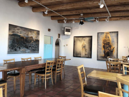 A cozy art gallery with wooden beams, featuring three paintings and a long wooden table surrounded by chairs.
