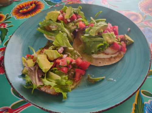 Three tostadas topped with lettuce, tomatoes, onions, and avocado on a colorful floral tablecloth.