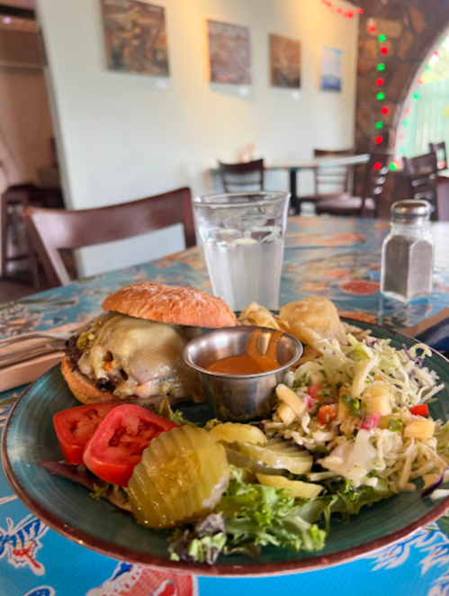 A plate with a cheeseburger, pickles, tomato, coleslaw, and a small cup of sauce, alongside a glass of water.