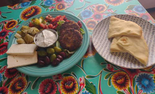 A colorful plate with a veggie patty, salad, olives, cheese, and a side of pita bread on a floral tablecloth.