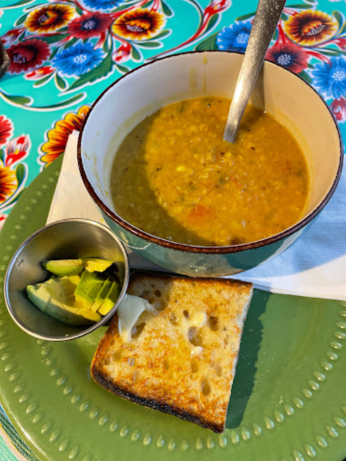 A bowl of soup with a spoon, a side of avocado slices, and a piece of toasted bread on a colorful floral tablecloth.