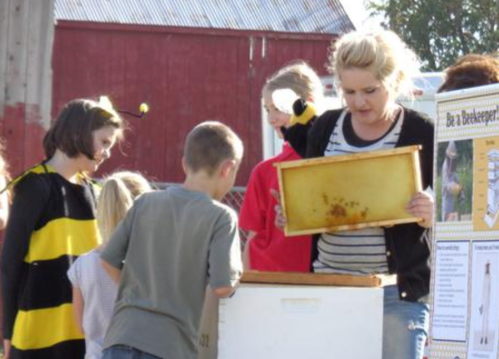 A woman shows a honeycomb frame to children at an outdoor event, with a red barn in the background.