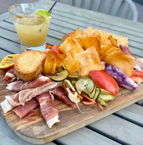 A wooden platter with assorted meats, vegetables, and crispy snacks, alongside a cocktail on a table.