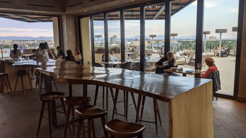 A modern bar with a curved wooden counter, overlooking a rooftop view with patrons enjoying drinks and meals.
