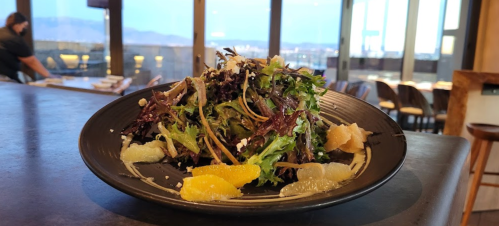 A vibrant salad with mixed greens and citrus segments, served on a black plate with a scenic view in the background.