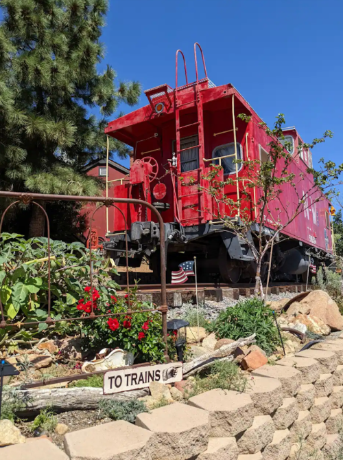 A red caboose sits in a garden with flowers and a sign pointing "TO TRAINS" under a clear blue sky.