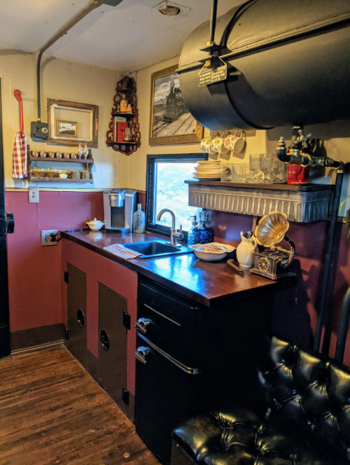 A cozy kitchen corner with a vintage sink, black fridge, and decorative items on shelves, featuring warm lighting.