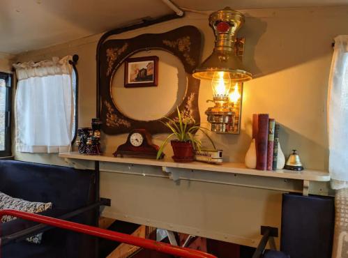 Cozy interior with a vintage lamp, framed photo, clock, books, and a potted plant on a shelf against a warm wall.