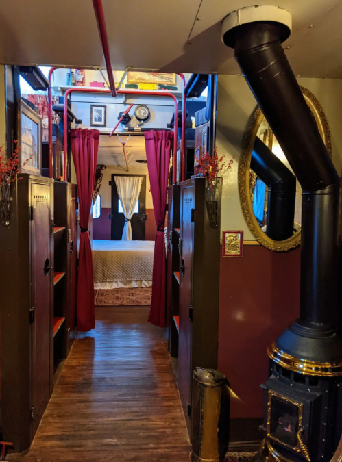 Cozy interior of a vintage-style room with a bed, bookshelves, and a wood stove, framed by red curtains.