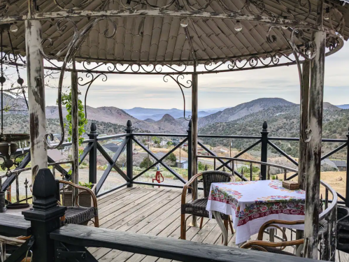 A scenic gazebo with a table and chairs overlooks rolling mountains and a valley under a cloudy sky.