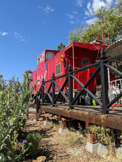 A bright red train caboose on a wooden platform surrounded by greenery and blue sky.