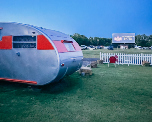 A vintage silver trailer parked on grass near a billboard advertising 89.7 FM, with cars in the background.