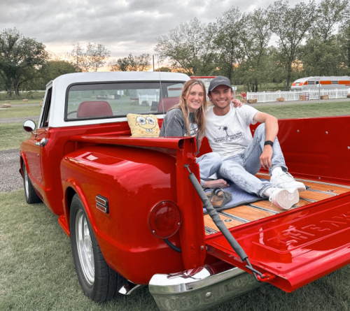 A couple sits in the back of a red vintage truck, smiling, with a SpongeBob pillow beside them and a scenic background.