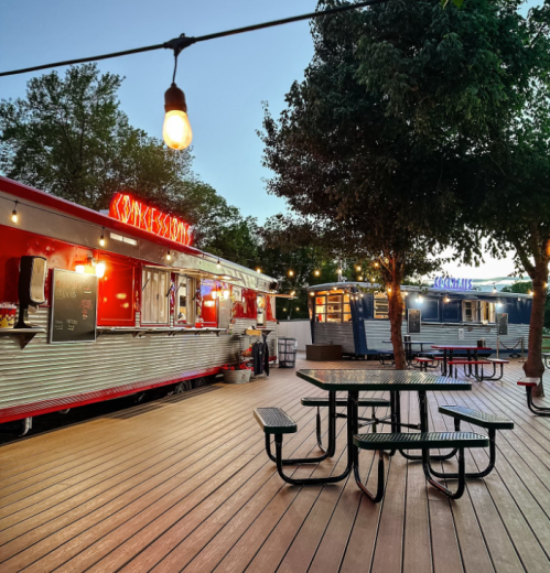 A vibrant food truck area with picnic tables, string lights, and trees, set against a twilight sky.