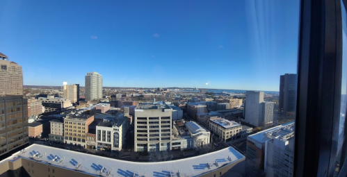 A panoramic view of a city skyline with buildings and a clear blue sky, overlooking a body of water in the distance.
