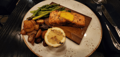 Grilled salmon with lemon, served on a wooden board, alongside asparagus and roasted potatoes on a white plate.