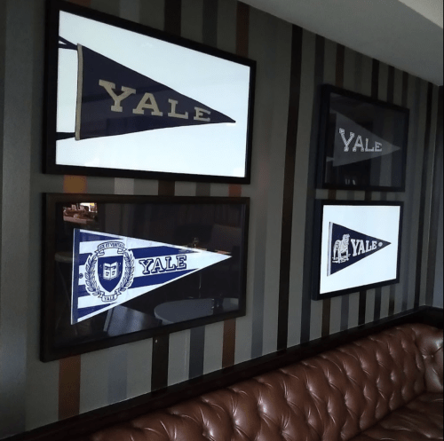 Framed Yale University pennants displayed on a striped wall above a brown leather couch.