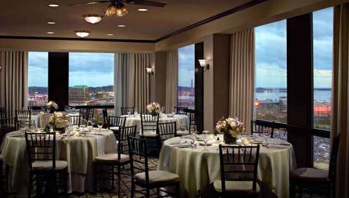 Elegant dining room with round tables set for a meal, large windows showcasing a city view at dusk.